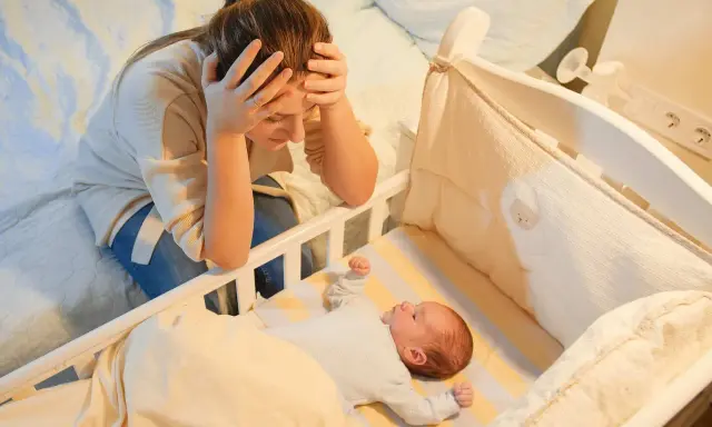 Madre agotada, con las manos en la cabeza, mira a su bebe que se desvela por la noche en su cuna.
