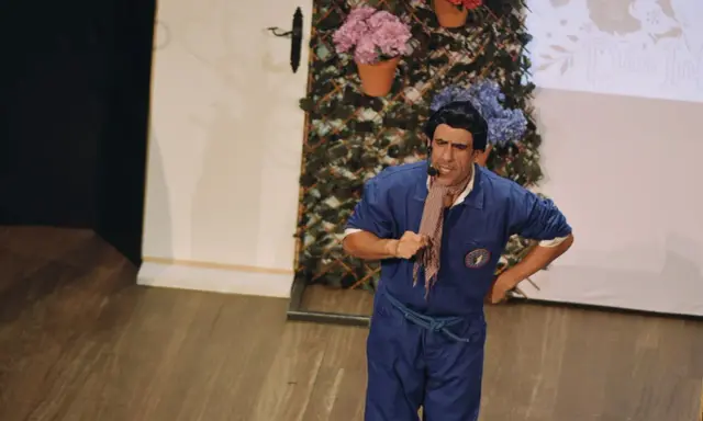 Actor con mono azul y pañuelo a cuadros, actuando en un teatro para niños. Fondo con flores y enredaderas.