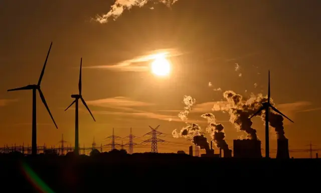 Silhouetten von Windrädern und rauchenden Industrieanlagen im Sonnenuntergang. Ein Bild, das zum Nachdenken über Stromkosten Norwegen vs. Deutschland anregt.