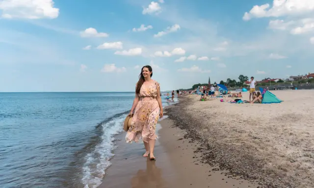 Kobieta w kwiecistej sukience spaceruje brzegiem morza w Sarbinowie. Na plaży widać ludzi odpoczywających na kocach i pod parasolami.