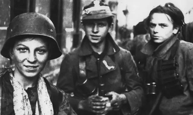 Trzech młodych żołnierzy, być może z Powstania Warszawskiego, w mundurach i hełmach. Jeden z nich uśmiecha się, gotowy do walki.