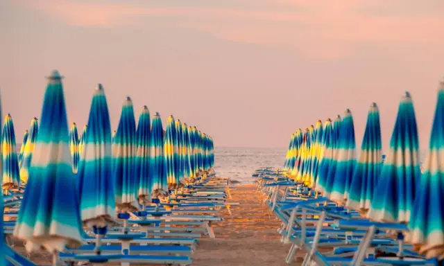Filetto di ombrelloni a righe blu e bianche sulla spiaggia, con il mare e il cielo rosa all'orizzonte. Un'immagine iconica della riviera adriatica.