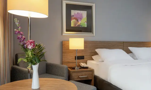 Ein Hotelzimmer mit Bett, Nachttischlampe, Sessel und Blumenstrauß.