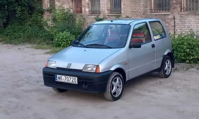 Srebrne cinquecento z czerwonymi akcentami we wnętrzu, zaparkowane na piaszczystym podwórku przed ceglanym budynkiem.