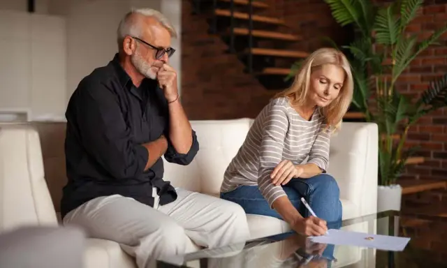 Un couple signe des documents, peut-être une désolidarisation de bail, dans un salon moderne. L'homme réfléchit, la femme écrit.