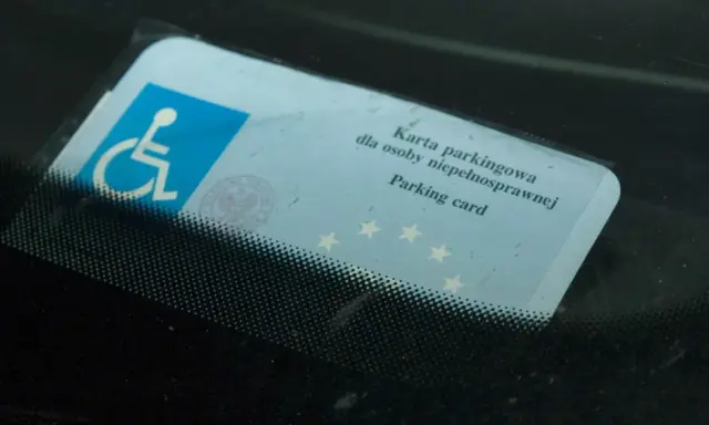 Niebieska karta parkingowa dla osoby niepełnosprawnej z symbolem wózka inwalidzkiego, umieszczona za przednią szybą samochodu.