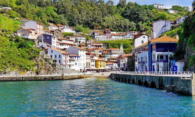 Bunte Häuser schmiegen sich an einen grünen Hang in Cudillero, Spanien. Das türkisfarbene Wasser des Hafens spiegelt die malerische Kulisse wider.