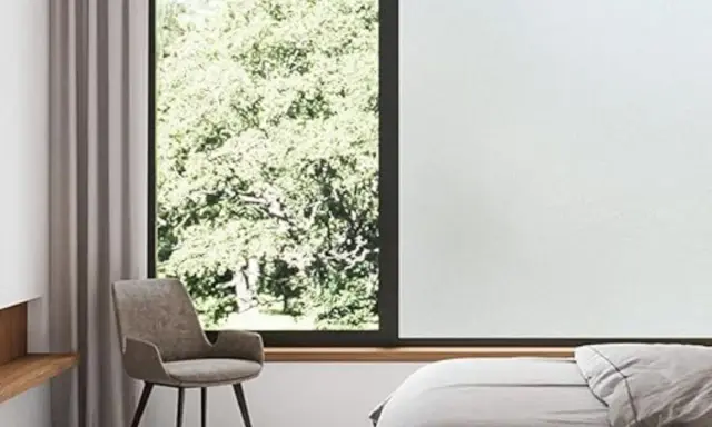 Habitación con cama, sillón y ventana con vidrio mate que filtra la luz del sol y la vegetación exterior.