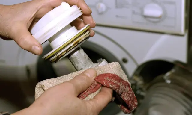 Hände reinigen das Flusensieb der Waschmaschine. Ein Tuch wischt Schmutz von dem ausgebauten Teil.