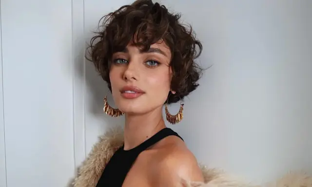 Mujer con un moderno corte shaggy rizado, ojos azules penetrantes y grandes pendientes dorados.