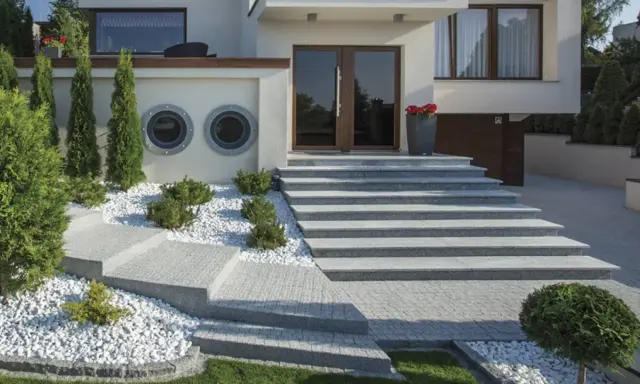 Nowoczesne schody zewnętrzne z beton dekoracyjny, otoczone białym żwirem i zielenią, prowadzące do drzwi wejściowych.