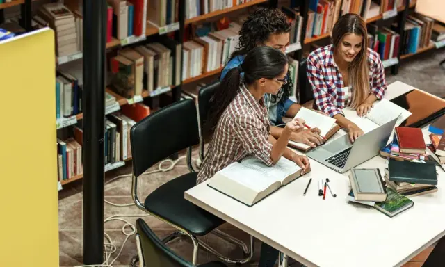 Trzy studentki pracują przy stole w bibliotece wyższej szkoły technicznej, otoczone książkami i laptopem.