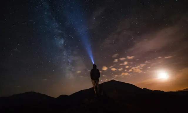 Una persona con una torcia illumina il cielo stellato su una montagna di notte. La Via Lattea è visibile.