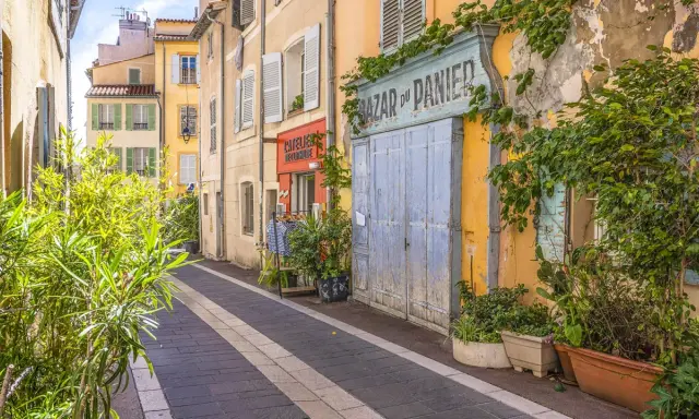 Bunte Häuser und Pflanzen säumen eine Gasse in der Marseille Altstadt. Ein "Bazar du Panier" Schild und blaue Holztüren fallen auf.
