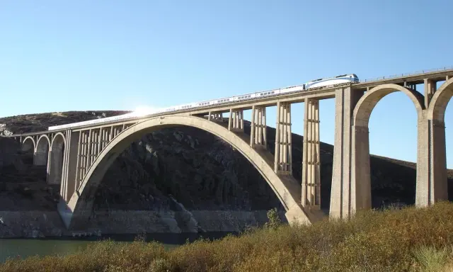 Un tren de alta velocidad cruza un imponente puente arco de hormigón sobre un embalse.