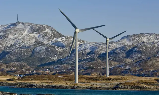 Zwei Windräder erzeugen sauberen Strom aus Norwegen. Im Hintergrund schneebedeckte Berge und ein kleines Dorf.