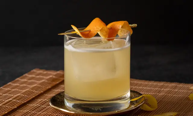 Ein erfrischender Rum Sour mit Eis und Orangenzesten garniert, serviert auf einem Bambusuntersetzer.