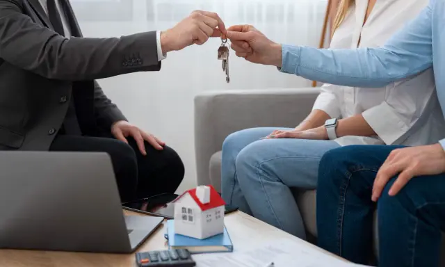 Agente inmobiliario entrega llaves a pareja. Un modelo de casa y calculadora sugieren una transacción, no una notificación de lanzamiento de desahucio.