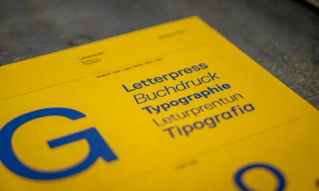 Żółta kartka z niebieskimi napisami: Letterpress, Buchdruck, Typographie, Leturpretun, Tipografia. Duża litera "G" po lewej. To praca typografa.