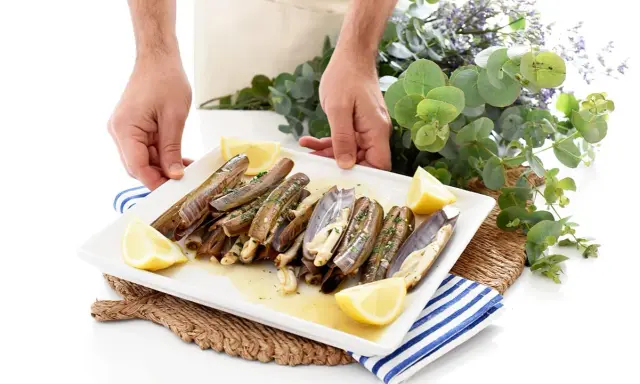 Manos sirviendo un plato de navajas a la plancha, decorado con gajos de limón y perejil picado.