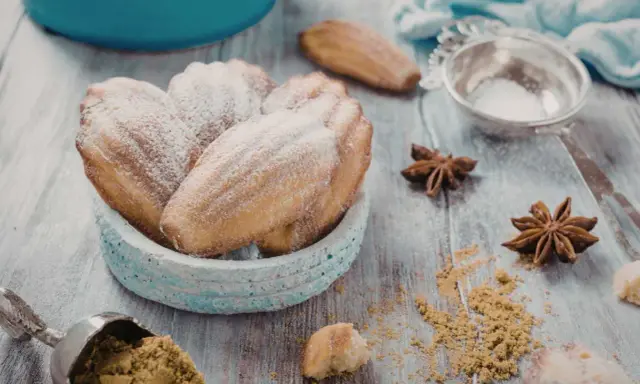 Un bol de biscuits madeleine noisette saupoudrés de sucre glace, avec des étoiles de badiane et une cuillère de poudre sur une table en bois.