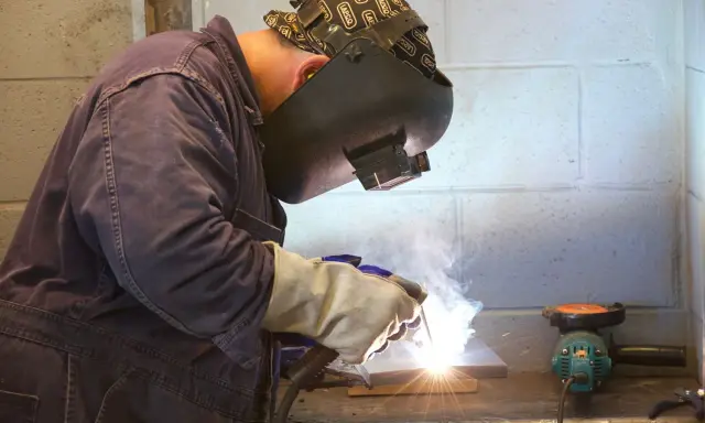 Soldador trabajando con un electrodo, creando chispas y humo. Se aprecian partes del electrodo en acción.