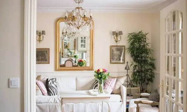 Un salotto con un divano bianco, un grande specchio dorato e un lampadario di cristallo, che evoca un elegante arredamento parigino.