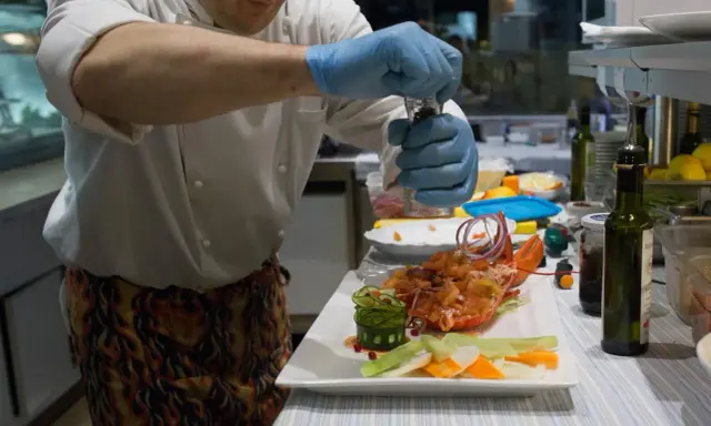Chef con guanti blu macina pepe su un'aragosta, un piatto che sembra uscito da "lo zodiaco Rimini".