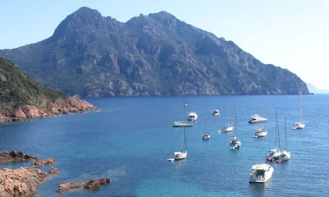 Vue d'une baie rocheuse avec plusieurs voiliers et bateaux à moteur en mouillage forain, sous une montagne imposante.