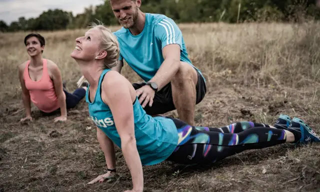 Ein Trainer unterstützt eine Frau bei einer Yoga-Übung im Freien. Das **Faszien Training** stärkt die Körpermitte.