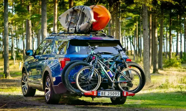 Niebieski SUV z dwoma rowerami na bagażniku i kajakami na dachu samochodu, gotowy na przygodę w lesie.