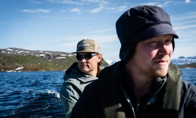 Två män i båt på en sjö, på väg mot fjällen. Solen skiner och det är en del av den stora matresan.
