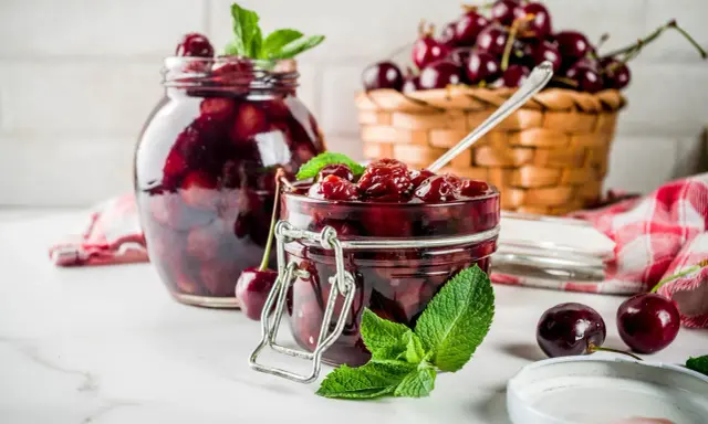 Słoiki pełne domowej konfitury z wiśni, ozdobione listkami mięty. Obok koszyk z dojrzałymi wiśniami i łyżeczka.
