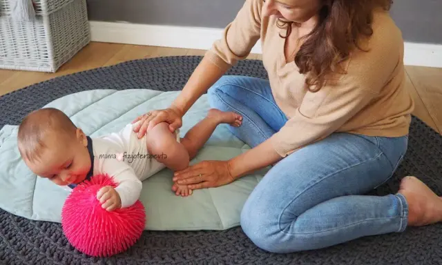 Fizjoterapeutka delikatnie masuje niemowlę, które leży na macie obok różowej piłeczki. Maluch, niczym mała amfibia, ćwiczy pod okiem mamy.