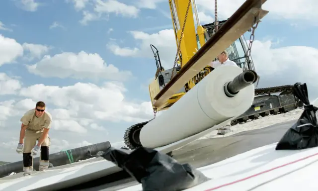Excavadora descarga un rollo de lámina bentonita. Obreros trabajan en la instalación bajo un cielo nublado.