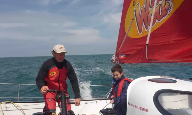 Franck-Yves Escoffier et un jeune garçon naviguent sur un voilier par temps ensoleillé.