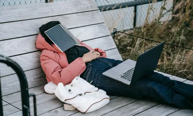 Osoba w różowej kurtce leży na ławce, z zeszytem na twarzy. Obok leży laptop i plecak. Czas na odpoczynek i dodanie nowych pomysłów.