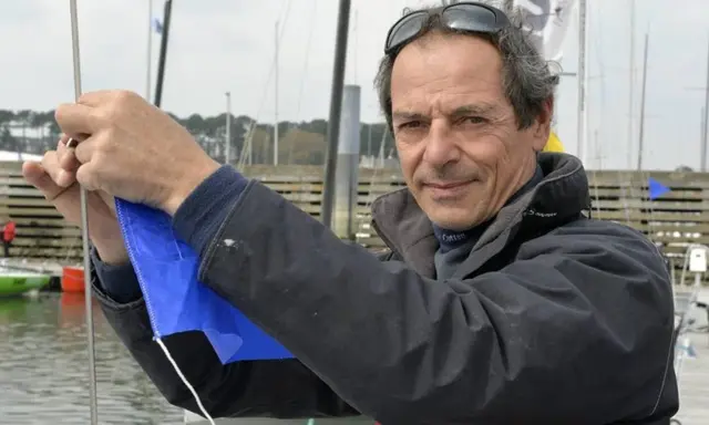 Marc Guillemot, skipper expérimenté, ajuste une voile bleue sur un ponton, prêt pour une nouvelle aventure en mer.