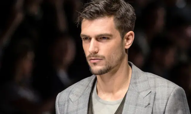 Uomo con capelli spettinati, sguardo intenso, indossa una giacca a quadri grigia.