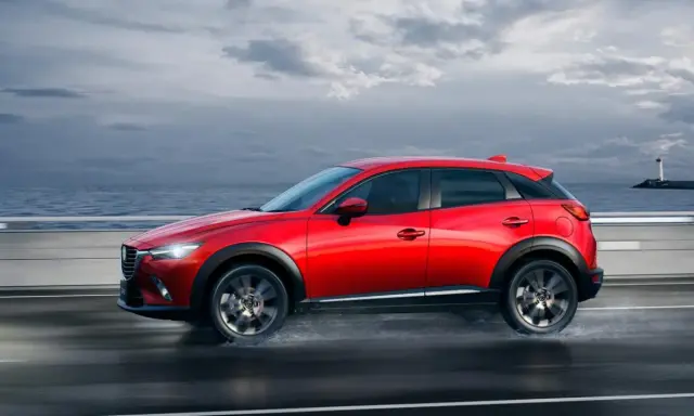 Un Mazda CX-3 rojo circula por una carretera mojada junto al mar. Las medidas del Mazda CX-3 son ideales para la ciudad y la aventura.