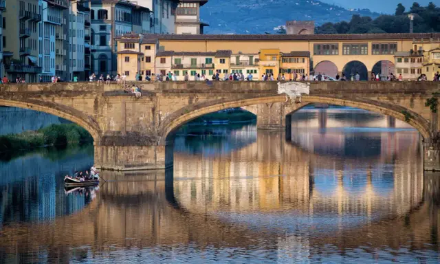 Le Ponte Vecchio à Florence, avec ses maisons colorées et ses reflets sur l'Arno. Une barque navigue paisiblement sous le pont.