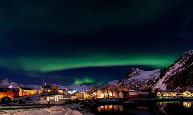 Lofoten norrsken över en snötäckt by med upplysta hus och speglingar i vattnet.