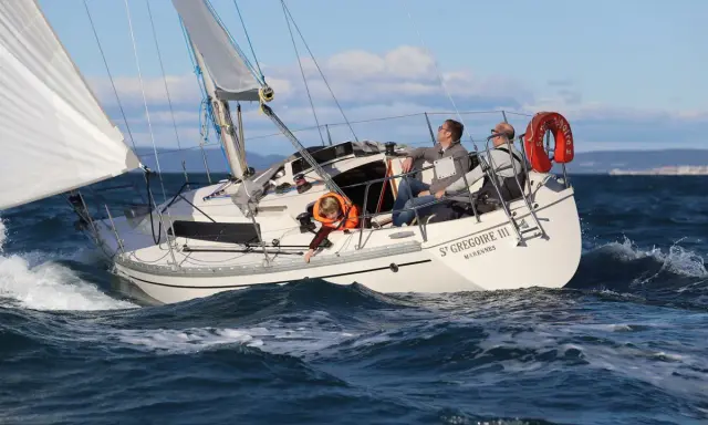 Un bateau symphonie navigue sur des eaux bleues, trois personnes à bord profitent du soleil.