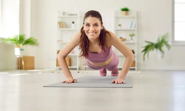 Frau in lila Sportkleidung macht Liegestütze für Yoga Muskelaufbau. Sie lächelt glücklich in die Kamera.
