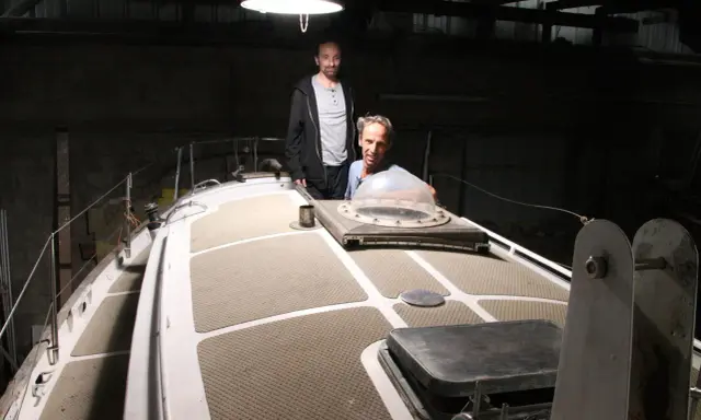 Nicole Van de Kerckhove et un autre homme sur le pont d'un voilier dans un hangar sombre.