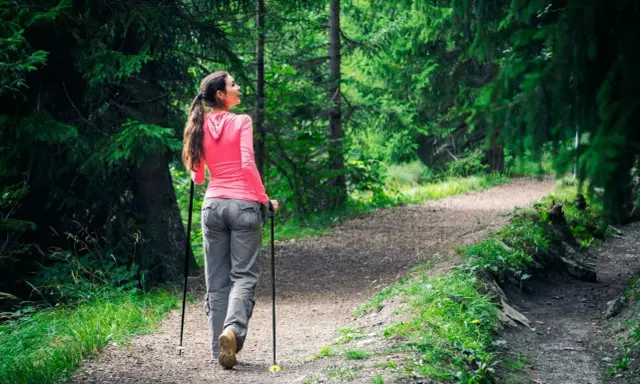 Kvinna går med stavar på en grusväg i en grönskande skog.