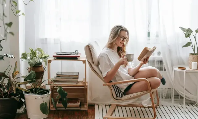 Mujer leyendo en un sillón, taza en mano, rodeada de plantas y libros. Un rincón de lectura con decoración acogedora y un tocadiscos.