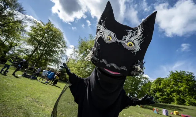 En stor, svart docka som liknar Syrsommarskuggan vinkar glatt i en park med folk i bakgrunden.