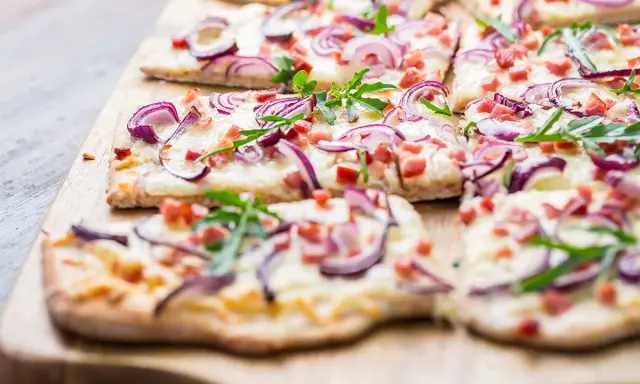 Flammkuchen-Ideen: Dünner Teig mit Käse, roten Zwiebeln, Speck und Rucola belegt. Perfekt für den Sommer!