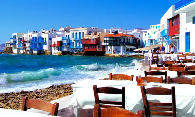 Cosa vedere a Mykonos in crociera: case colorate sul mare, onde che si infrangono sulla spiaggia di ciottoli e tavoli apparecchiati per godersi il panorama.